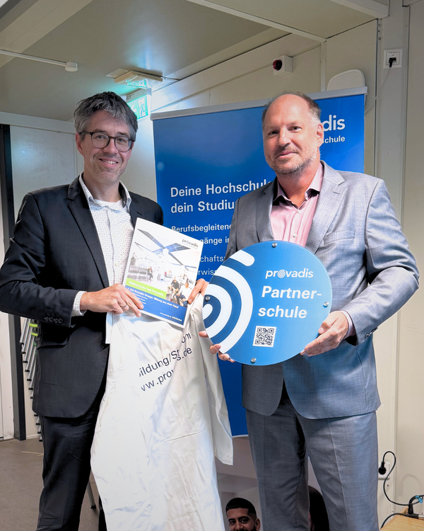 Prof. Björn Hekman, Hochschulvorstand der Provadis Hochschule, hält ein Schild mit der Aufschrift „Provadis Partnerschule“. Neben ihm steht eine weitere Person mit einem Dokument und einer Tasche. Im Hintergrund ist ein blaues Provadis-Banner zu sehen. Anlass ist die Vertragsunterzeichnung: Das Gymnasium Süd wird offizielle Partnerschule der Provadis Hochschule.